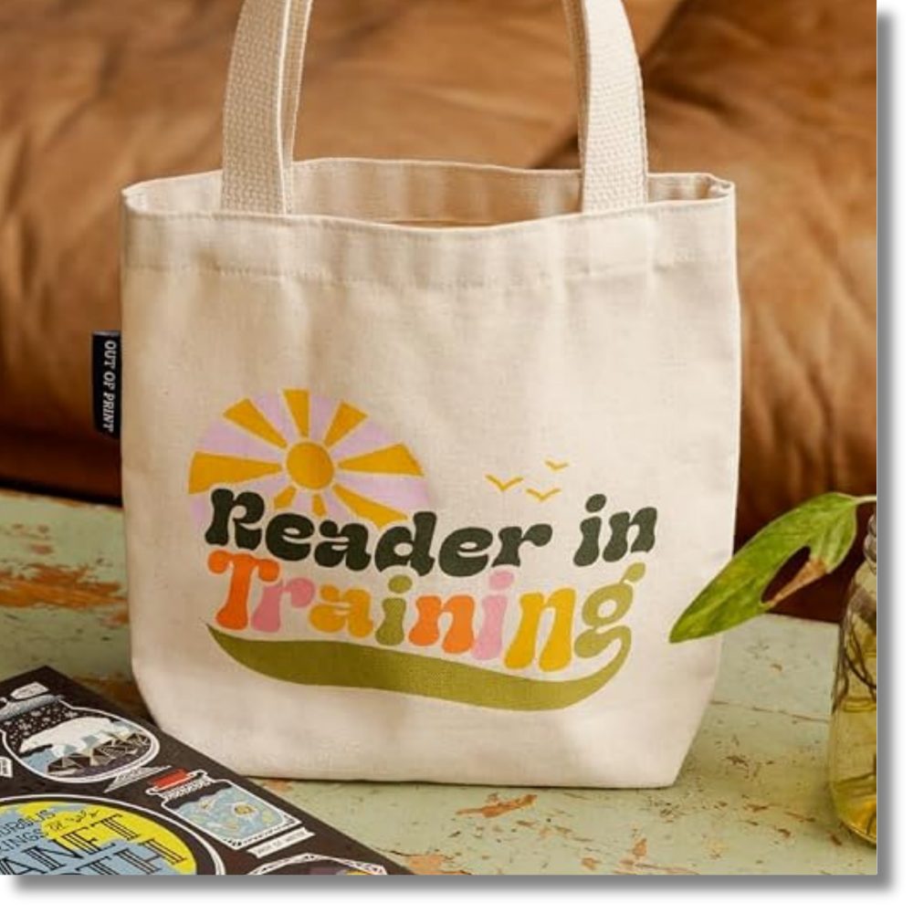 A cream canvas tote bag with the words 'Reader in Training' printed in a retro multicolor font, featuring a yellow and orange sun graphic. The bag is styled on a rustic table with a book and a small plant nearby.