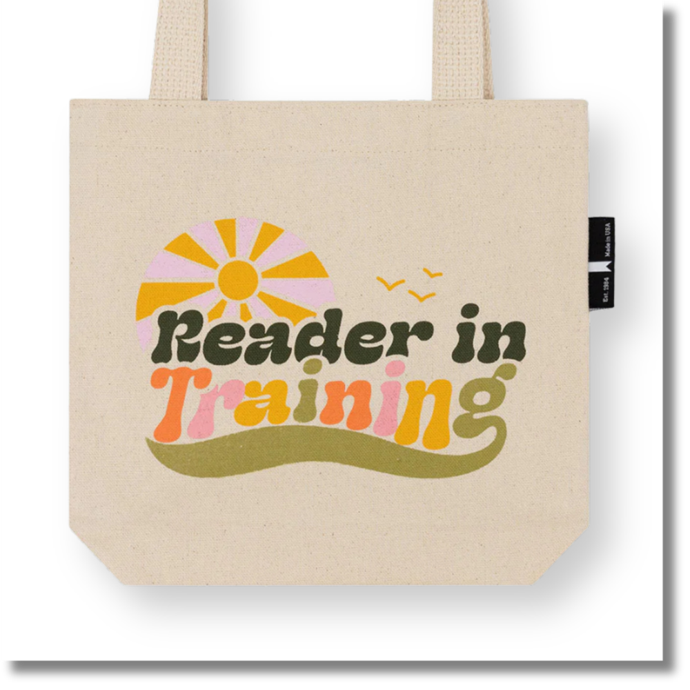 A beige canvas tote bag with a retro-style graphic that reads 'Reader in Training' in dark green and pastel pink, orange, and yellow lettering, under a pink and yellow sun and two small birds. A black side tag shows the brand 'The Paper Bag Company'.